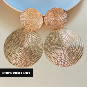 May include: Gold-toned, textured disc earrings. Each earring features a smaller, circular top piece and a larger, circular bottom piece. The earrings have a detailed, patterned surface. The text "SHIPS NEXT DAY" is visible in a black rectangle.