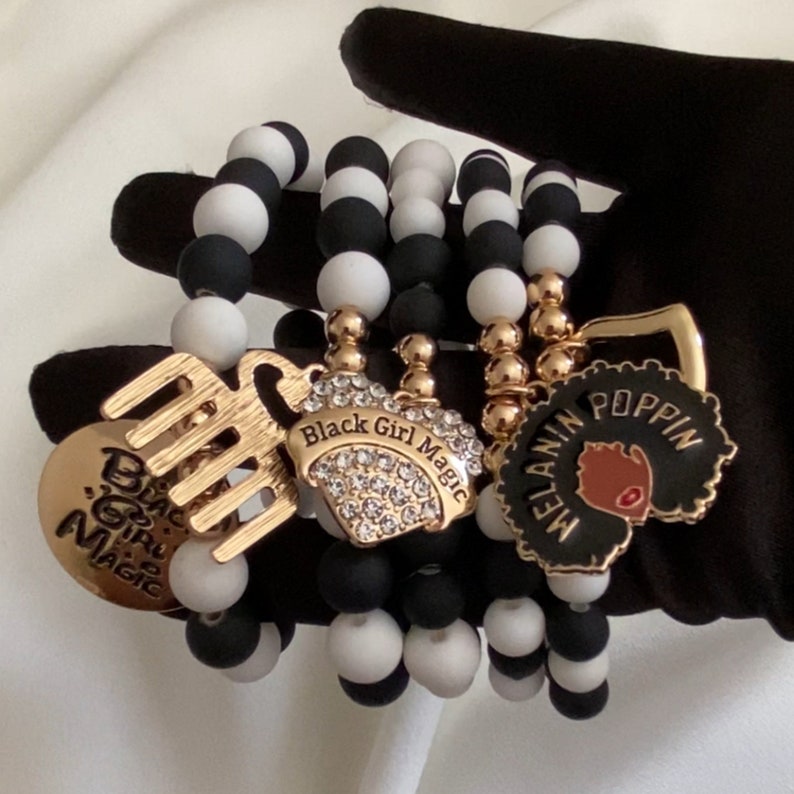 May include: A set of beaded bracelets in black and white. Each bracelet features gold charms with the phrases "Black Girl Magic" and "Melanin Poppin". The bracelets are a stylish accessory.