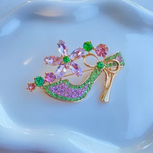 Op de afbeelding: Een goudkleurige broche in de vorm van een high-heelschoen, versierd met roze, groene en paarse kristallen. De schoen heeft een bloemenontwerp en een strik, wat een glamoureus accessoire creëert. De broche ligt op een wit oppervlak.