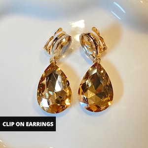 Può includere: Orecchini a clip con grandi cristalli a goccia color ambra. Gli orecchini hanno dettagli in metallo color oro e un meccanismo a clip. Il testo "CLIP ON EARRINGS" è visualizzato in un riquadro nero in basso.