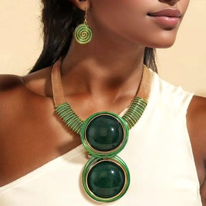 May include: A green and gold necklace and earring set. The necklace features two large, round, dark green pendants with gold trim. The necklace and earrings have a spiral design with green and gold accents. The jewelry is worn on a person wearing a white top.