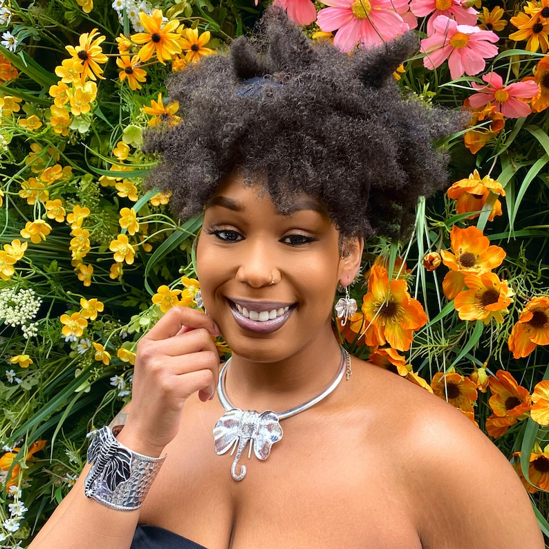 May include: A silver elephant pendant necklace, matching earrings, and a cuff bracelet. The jewelry features intricate details and is set against a backdrop of colorful flowers. The elephant pendant has a rhinestone design.