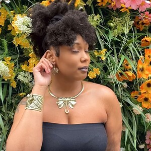 May include: A woman with dark curly hair wearing a black strapless top and gold jewelry. The jewelry includes a large elephant-shaped pendant necklace and a matching pair of earrings. The woman is standing in front of a wall of flowers.