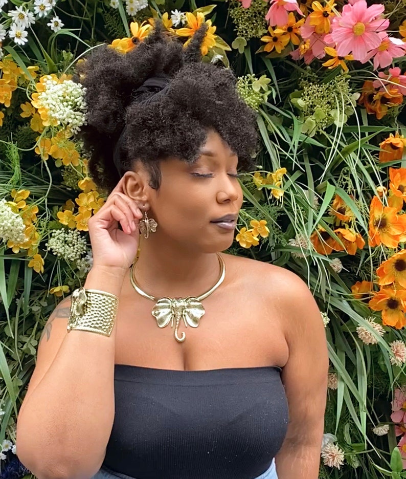 May include: A woman with dark curly hair wearing a black strapless top and gold jewelry. The jewelry includes a large elephant-shaped pendant necklace and a matching pair of earrings. The woman is standing in front of a wall of flowers.