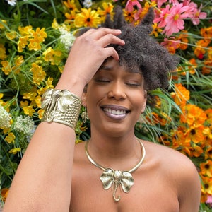 May include: A woman with dark curly hair is wearing a gold elephant pendant necklace and a gold elephant cuff bracelet. She is smiling and has her hand on her head. The background is a wall of yellow flowers.