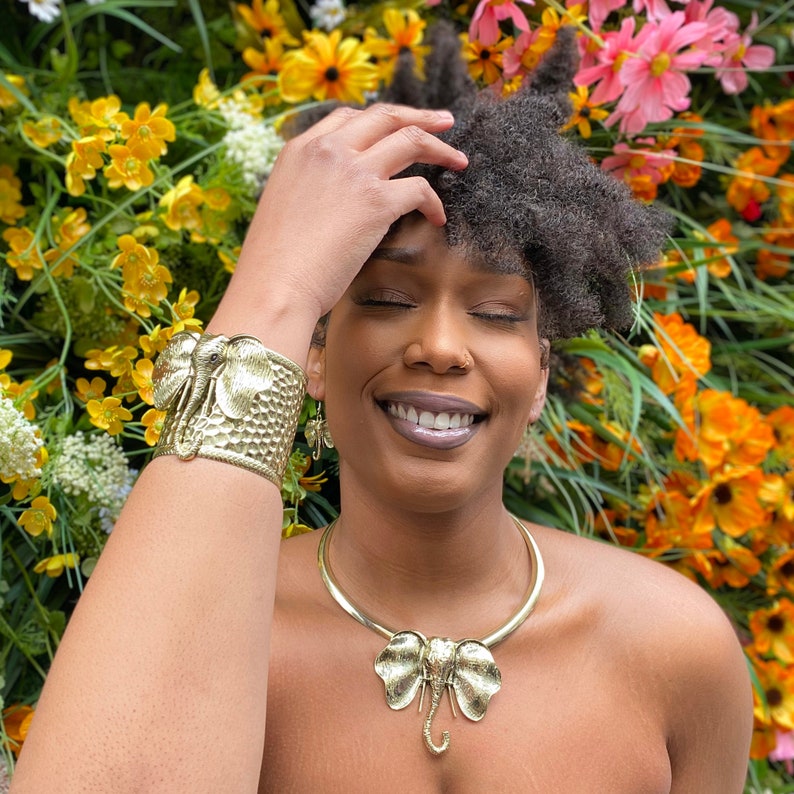May include: A woman with dark curly hair is wearing a gold elephant pendant necklace and a gold elephant cuff bracelet. She is smiling and has her hand on her head. The background is a wall of yellow flowers.