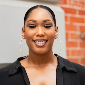 May include: A woman with dark brown skin is smiling and looking directly at the camera. She is wearing a black collared shirt and large silver hoop earrings. The background is a brick wall.