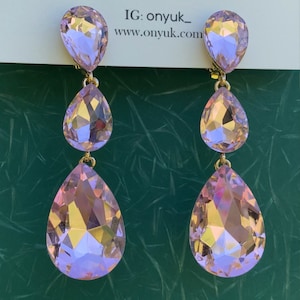 May include: A pair of elegant, three-tiered drop earrings. Each earring features three iridescent, teardrop-shaped crystals in shades of pink and yellow, connected by gold-colored metal. The earrings are clipped to the ear. The text "IG: onyuk_ www.onyuk.com" is visible.