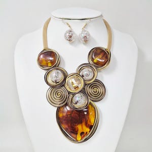 Pode incluir: Um conjunto de colar e brincos exibido em um manequim branco. O colar apresenta grandes pedras de cor âmbar e detalhes enrolados em arame. Os brincos são pequenos, transparentes e esféricos.
