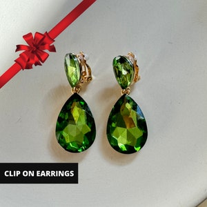 May include: A pair of clip-on earrings with a gold-tone setting. Each earring features a faceted, pear-shaped green gemstone suspended below a smaller, oval-shaped green gemstone. The text "CLIP ON EARRINGS" is displayed in a white box.