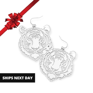 May include: A pair of white, laser-cut tiger head earrings with silver-colored ear wires. The earrings are detailed with intricate cut-out patterns. A red ribbon and bow are in the upper left corner. The text "SHIPS NEXT DAY" is in a black rectangle.
