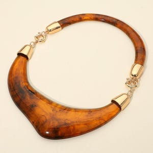 May include: A striking necklace with a tortoiseshell-patterned, curved pendant. It has gold-coloured metal details and a clasp. The warm brown and amber hues create a bold, eye-catching accessory, perfect for adding a touch of glamour.