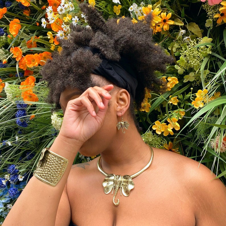 May include: A person wearing a gold elephant pendant necklace and gold earrings. The person is wearing a black headband and has dark hair. The background is a wall of green leaves and flowers.