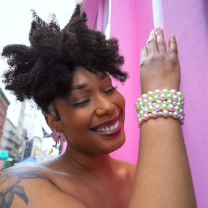 May include: A woman wearing a pink and green pearl bracelet and pink elephant earrings. The woman is smiling with her eyes closed. The background is a bright pink wall.