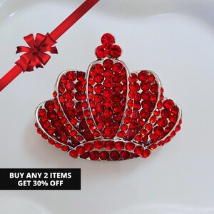 May include: A silver-toned crown brooch, encrusted with numerous red rhinestones. The crown design is detailed and ornate, with a classic royal shape. The image also includes a red ribbon and the text "BUY ANY 2 ITEMS GET 30% OFF".