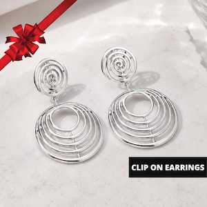 May include: A pair of silver-toned clip-on earrings featuring a small spiral stud connected to a larger circular drop component. The drop consists of four concentric rings of varying sizes. The text CLIP ON EARRINGS is visible in the corner.