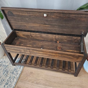May include: A dark brown wooden bench with a hinged lid that opens to reveal a storage compartment. The bench has a black metal frame and slatted shelves underneath.