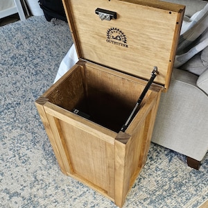May include: A wooden storage box with a hinged lid and a spring-loaded mechanism. The box is made of natural wood and has a light brown finish. The lid has a logo that says "Tinney Outfitter".