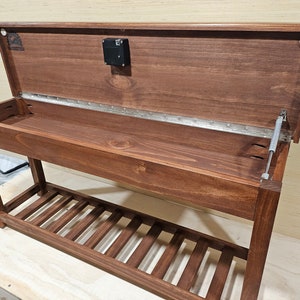 May include: A wooden bench with a hinged top that opens to reveal storage space. The bench has a lower shelf with slatted supports. The bench is stained a dark brown color.