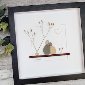 May include: A framed pebble art piece depicting a couple sitting on a branch with a heart above them. The artwork is on a white background with brown twigs and a brown branch.