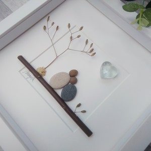 May include: A white framed picture with a twig, dried flowers, and a heart-shaped glass piece. The twig is decorated with small stones and the word "Forever" is written on the twig.