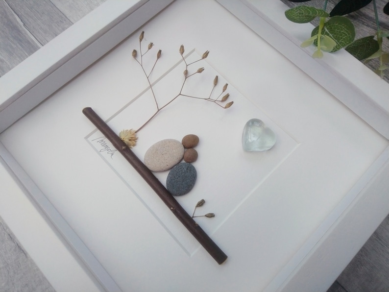 May include: A white framed picture with a twig, dried flowers, and a heart-shaped glass piece. The twig is decorated with small stones and the word "Forever" is written on the twig.
