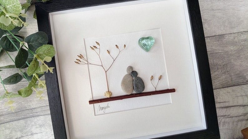 May include: A framed pebble art piece depicting two figures holding each other, sitting on a branch under a tree with a small green heart above them. The piece is signed "Manjula".