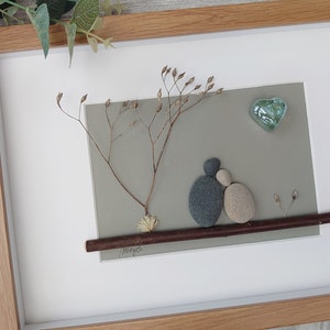 May include: A framed pebble art piece depicting a couple sitting on a branch under a tree with a heart-shaped glass ornament above them. The background is a light gray color and the branch is brown. The couple is made of gray and beige pebbles.