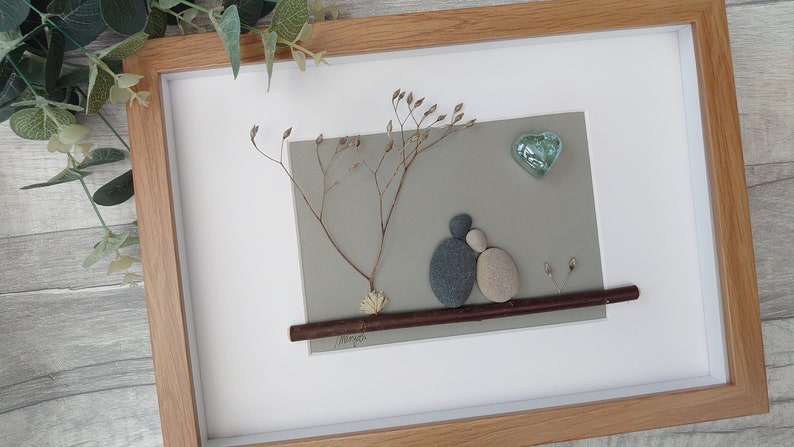 May include: A framed pebble art piece depicting a couple sitting on a branch under a tree with a heart-shaped glass ornament above them. The background is a light gray color and the branch is brown. The couple is made of gray and beige pebbles.