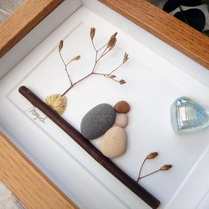 May include: A framed artwork featuring a small arrangement of stones, a twig, and dried flowers. The stones are arranged to look like a bird. A blue glass heart is in the upper right corner. The artwork is signed "Magnolia".
