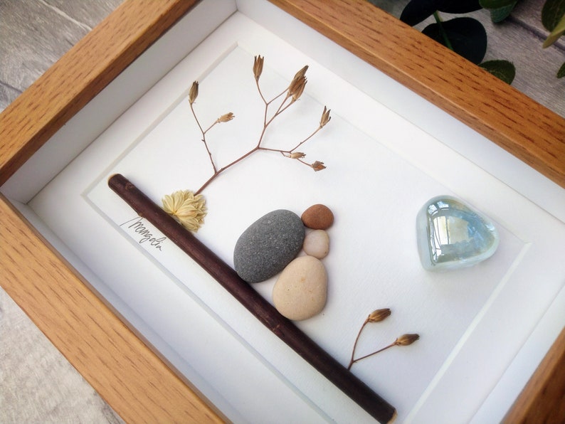 May include: A framed artwork featuring a small arrangement of stones, a twig, and dried flowers. The stones are arranged to look like a bird. A blue glass heart is in the upper right corner. The artwork is signed "Magnolia".