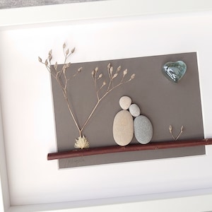 May include: A framed pebble art piece depicting two figures sitting on a branch with a heart-shaped glass piece above them. The background is a gray paper with dried flowers.