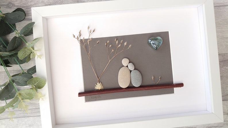 May include: A framed pebble art piece depicting two figures sitting on a branch with a heart-shaped glass piece above them. The background is a gray paper with dried flowers.