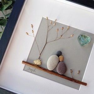 May include: A framed pebble art piece depicting two figures sitting on a branch with a heart-shaped piece of glass above them. The background is a light gray color and the branch is brown. The piece is signed "Mayah" in the bottom left corner.