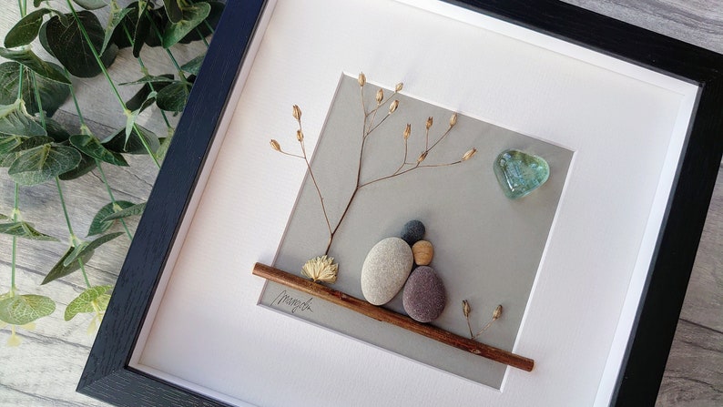 May include: A framed pebble art piece depicting two figures sitting on a branch with a heart-shaped piece of glass above them. The background is a light gray color and the branch is brown. The piece is signed "Mayah" in the bottom left corner.