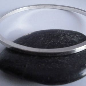 May include: A silver bangle bracelet with a smooth, polished finish. The bracelet is resting on a dark grey stone.
