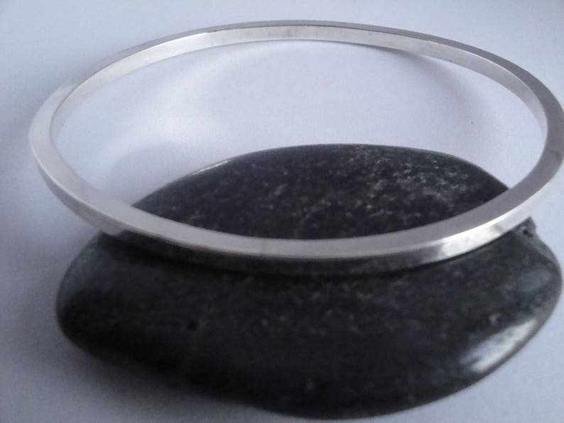 May include: A silver bangle bracelet with a smooth, polished finish. The bracelet is resting on a dark grey stone.