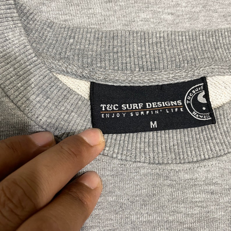 May include: Close-up of a gray sweatshirt with a crew neck. The black label reads "T&C Surf Designs Enjoy Surfin' Life" with a yin-yang symbol and the word "Hawaii". The size "M" is also visible.