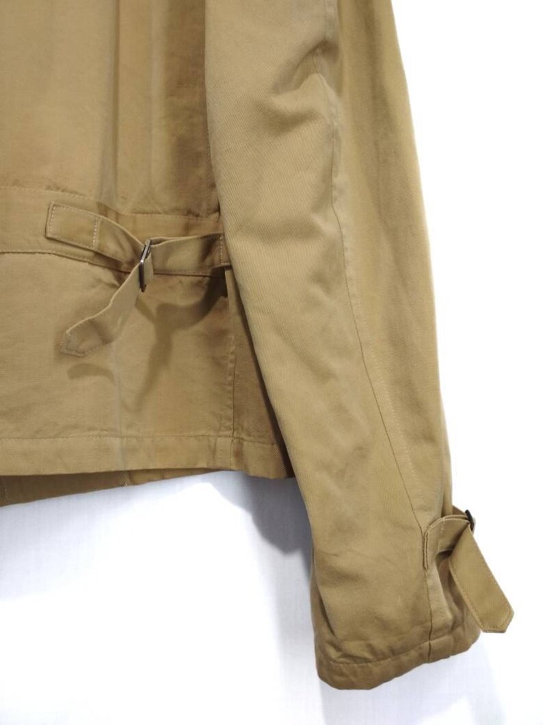May include: A tan utility jacket with a belt and buckle closure. The jacket has a long sleeve with a buttoned cuff.