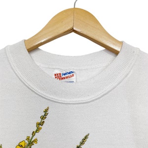 Op de afbeelding: Witte Hanes Activewear sweatshirt met een gele bloemenprint. Het label zegt "Hanes Activewear".