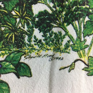 Op de afbeelding: Witte stof met een groene bloemenprint. De stof heeft een textuur en de print toont verschillende soorten bladeren en planten.
