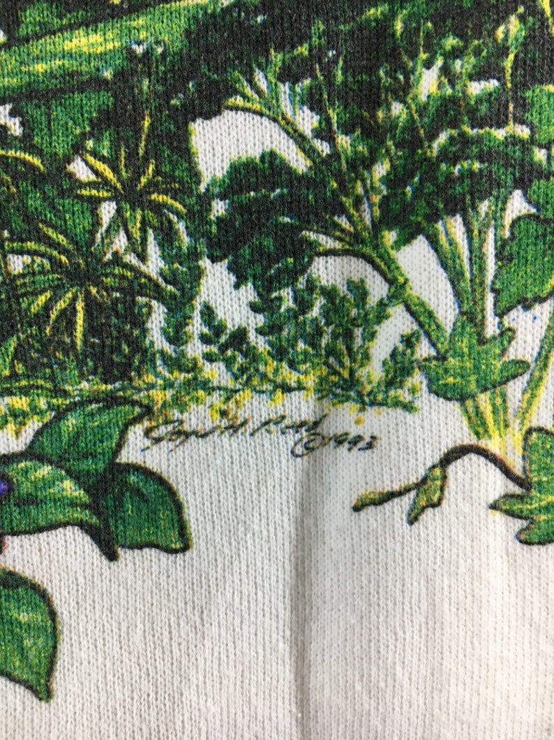 Op de afbeelding: Witte stof met een groene bloemenprint. De stof heeft een textuur en de print toont verschillende soorten bladeren en planten.