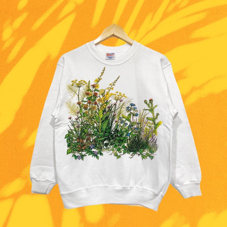 Op de afbeelding: Witte trui met een kleurrijke bloemenprint van wilde bloemen en groen. De print is gecentreerd op de voorkant van de trui.