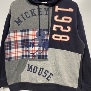 Puede incluir: Una sudadera con capucha de color azul, gris y rojo con un gr&aacute;fico de Mickey Mouse y el texto "Mickey Mouse" y "1928".