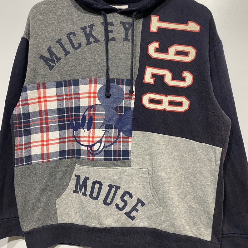 Puede incluir: Una sudadera con capucha de color azul, gris y rojo con un gr&aacute;fico de Mickey Mouse y el texto "Mickey Mouse" y "1928".