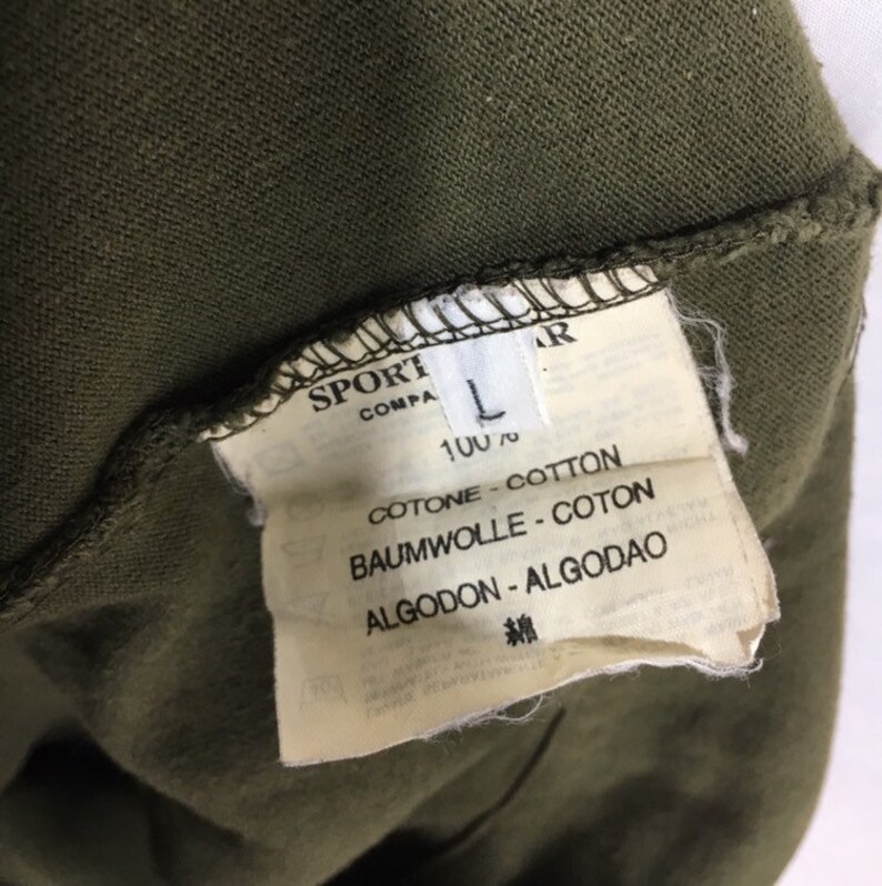 May include: A white tag with black text that reads "SPORT COMPA L 100% COTONE - COTTON BAUMWOLLE - COTON ALGODON - ALGODAO 綿"