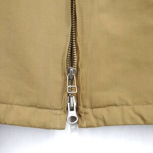 May include: Close-up of a zipper on a brown jacket. The zipper is silver and has a pull tab.