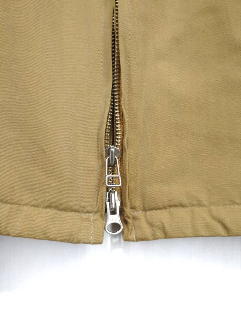 May include: Close-up of a zipper on a brown jacket. The zipper is silver and has a pull tab.