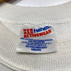 Op de afbeelding: Wit Hanes Activewear-etiket met rode letters. Het etiket vermeldt "50% Katoen (Coton/Algodon), 50% Polyester, Gemaakt in de VS Fabriqu&eacute; aux E-U Hecho en los E.U.A., Moyen/Mediano Medium."
