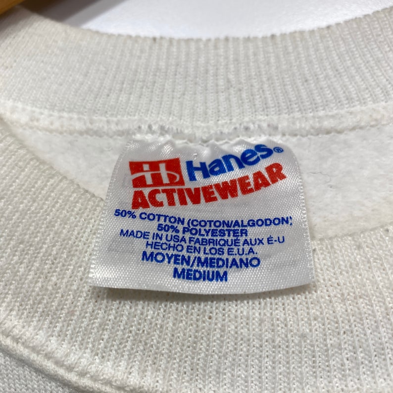 Op de afbeelding: Wit Hanes Activewear-etiket met rode letters. Het etiket vermeldt "50% Katoen (Coton/Algodon), 50% Polyester, Gemaakt in de VS Fabriqu&eacute; aux E-U Hecho en los E.U.A., Moyen/Mediano Medium."